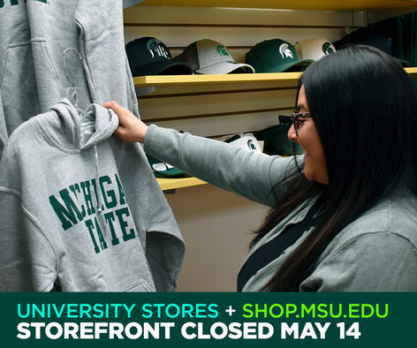 Woman holds and examines a gray Michigan State hoodie in a storefront, surrounded by other Michigan State apparel and accessories.