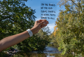 A person holding up a clear sign in front of the Red Cedar River. On the sign reads "On the banks of the Red Cedar, there's a school that's known to all."